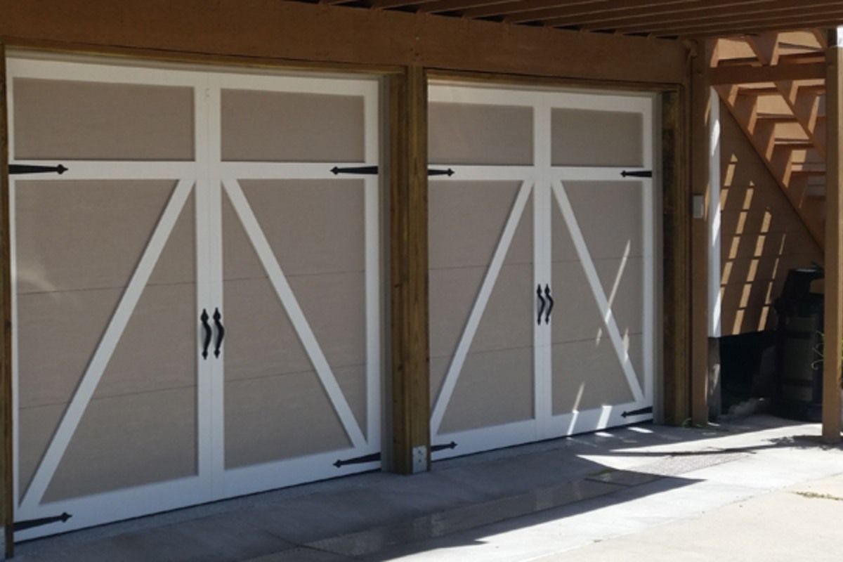 Two beige garage doors with white trim, black handles and hinges under a wooden structure.