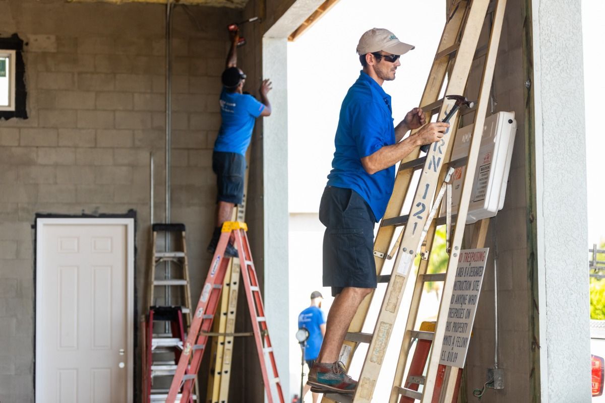 Two men, wearing blue shirts, work on construction. One on a ladder, the other on a taller ladder, both near a concrete wall.