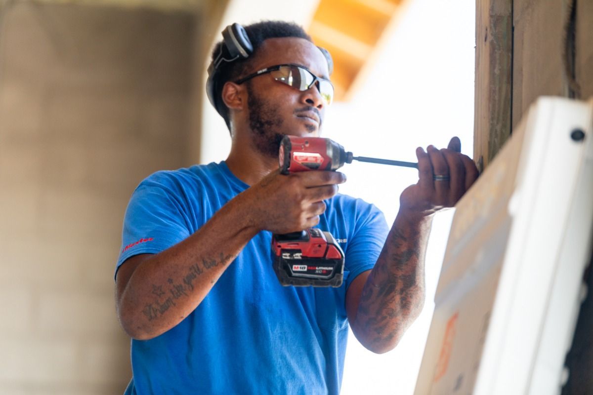 Man in blue shirt using a power drill. He wears safety glasses and ear protection, working inside a building.