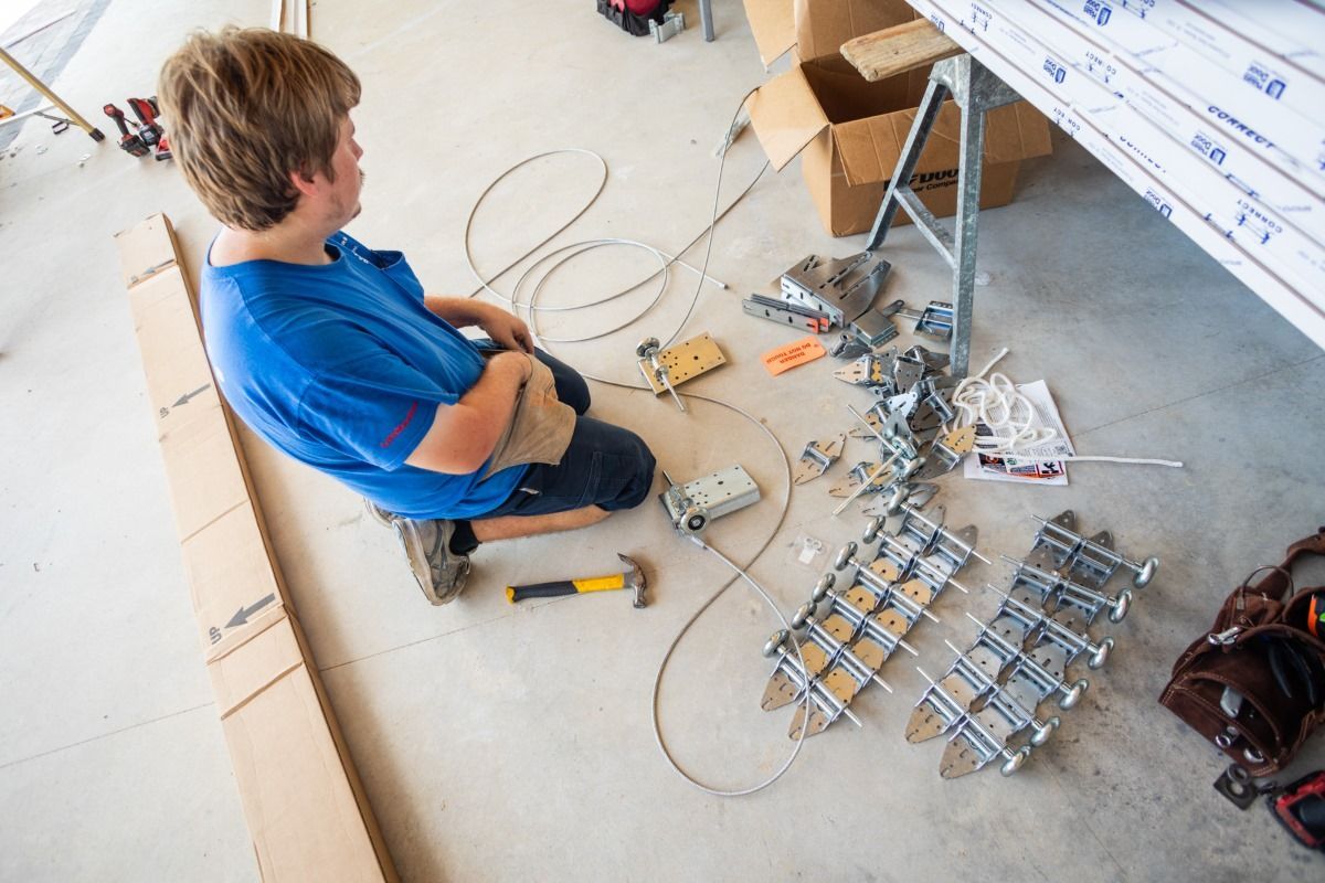 Person kneeling on floor, assembling parts with a hammer. Tools, boxes, and equipment scattered around.