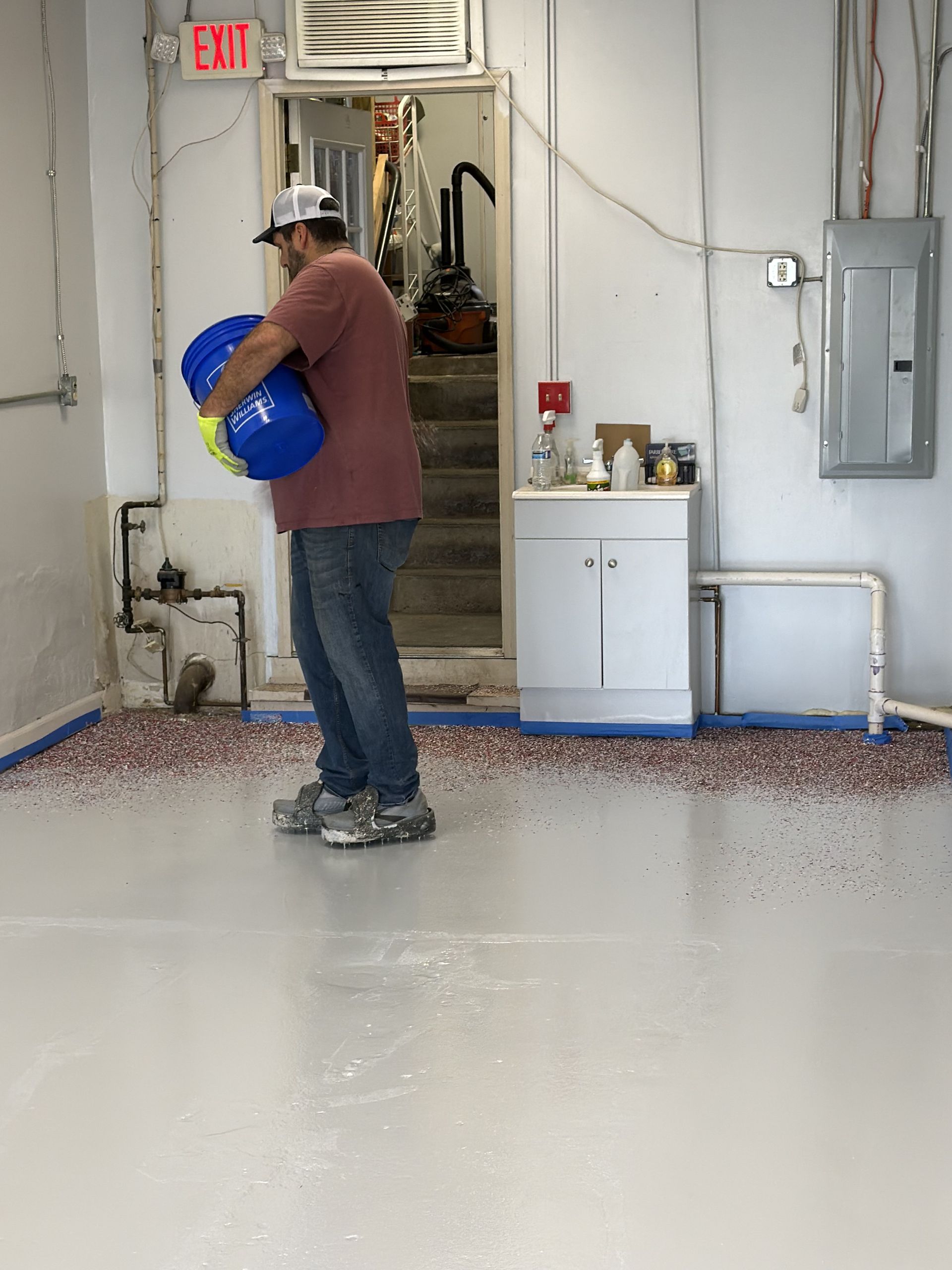 A worker holding a blue bucket spreads colorful decorative flakes onto a freshly coated gray epoxy floor in an interior.