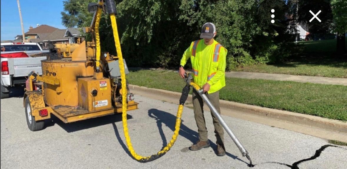 A worker in a high-visibility vest uses a heated sealant machine to repair a crack in an asphalt road.