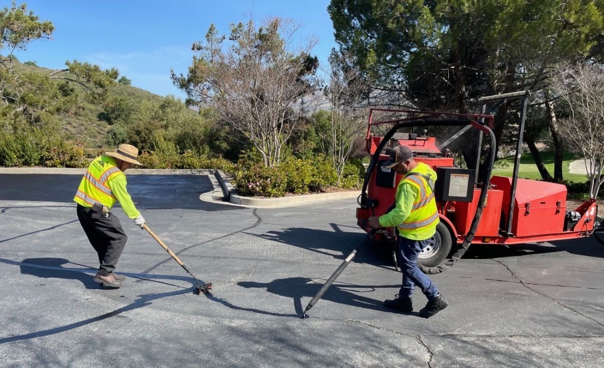 Two workers in neon safety vests use equipment to seal cracks in a paved parking lot on a sunny day.