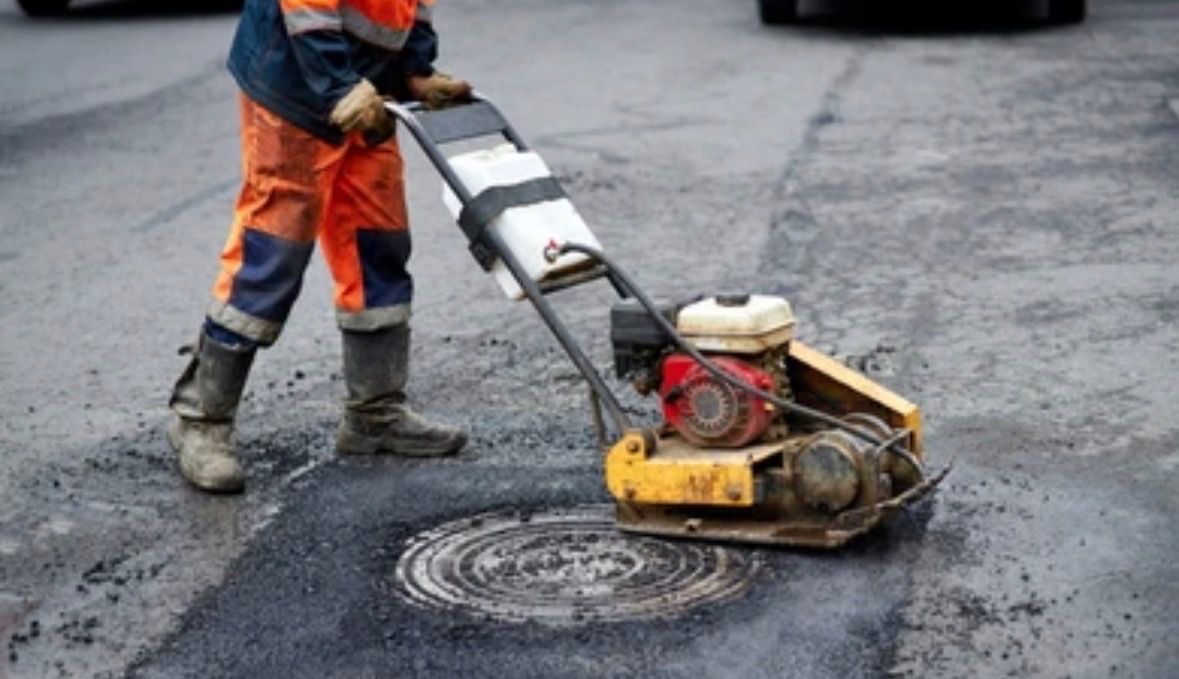 A road worker uses a vibratory plate compactor to smooth asphalt around a circular manhole cover on a street.