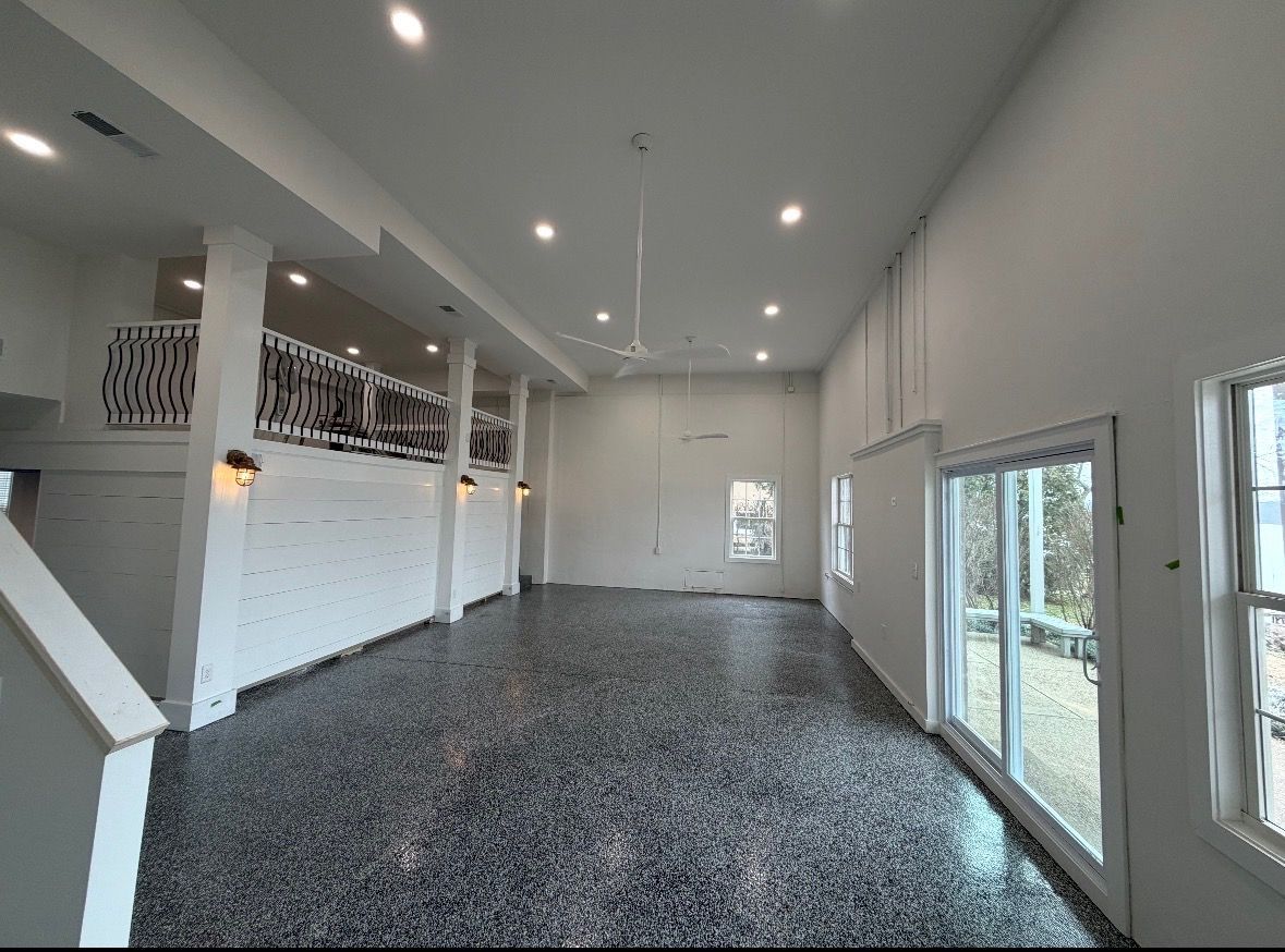 A spacious, white-walled room with high ceilings, recessed lighting, a decorative loft railing, and dark speckled flooring.
