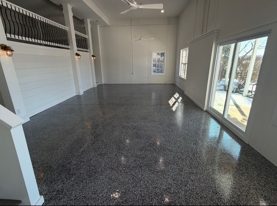 A spacious, high-ceilinged room with glossy speckled flooring, a ceiling fan, white walls, and glass sliding doors.