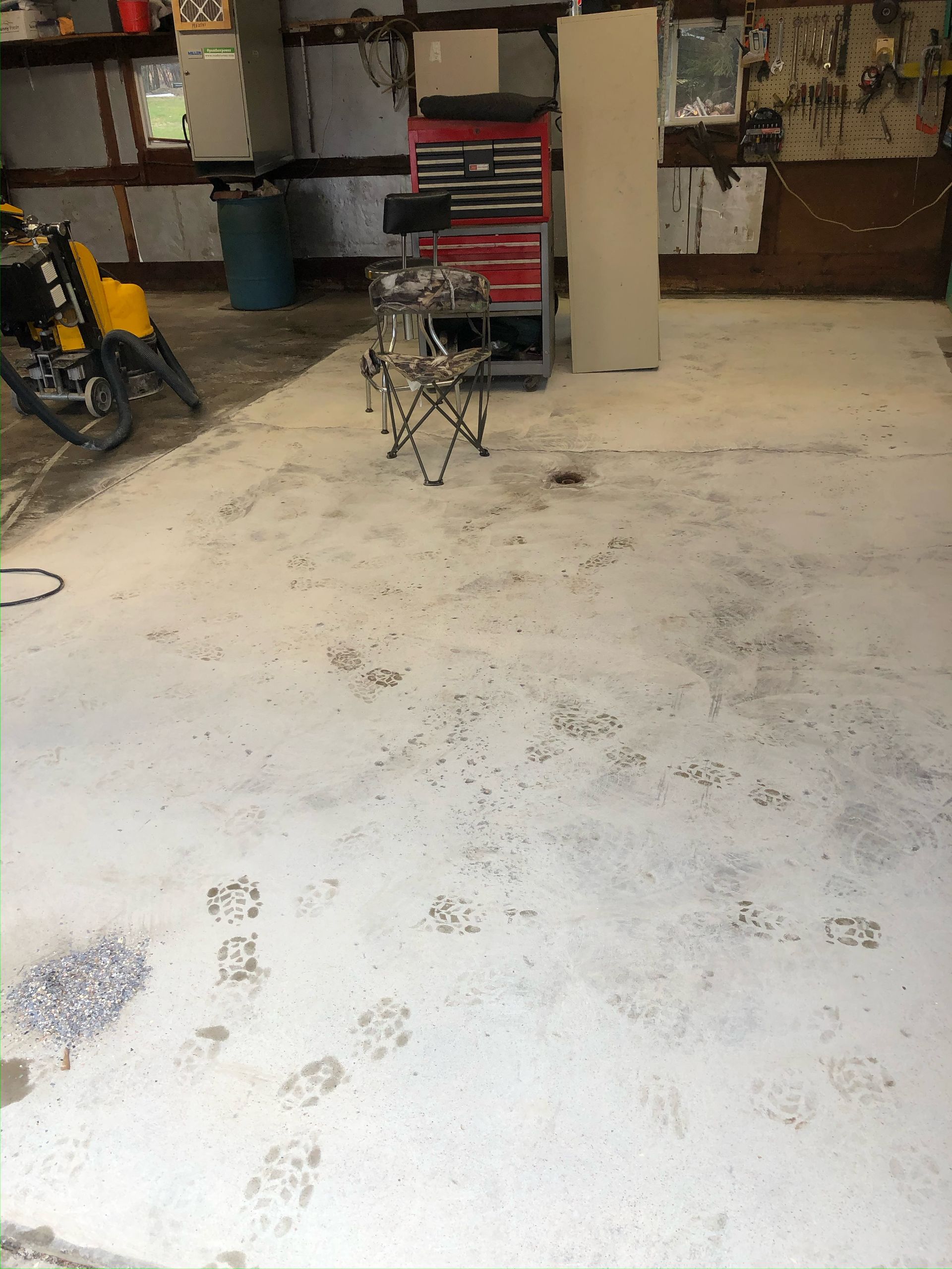A garage floor with white painted concrete featuring dark muddy footprints leading toward a metal tool chest and chair.