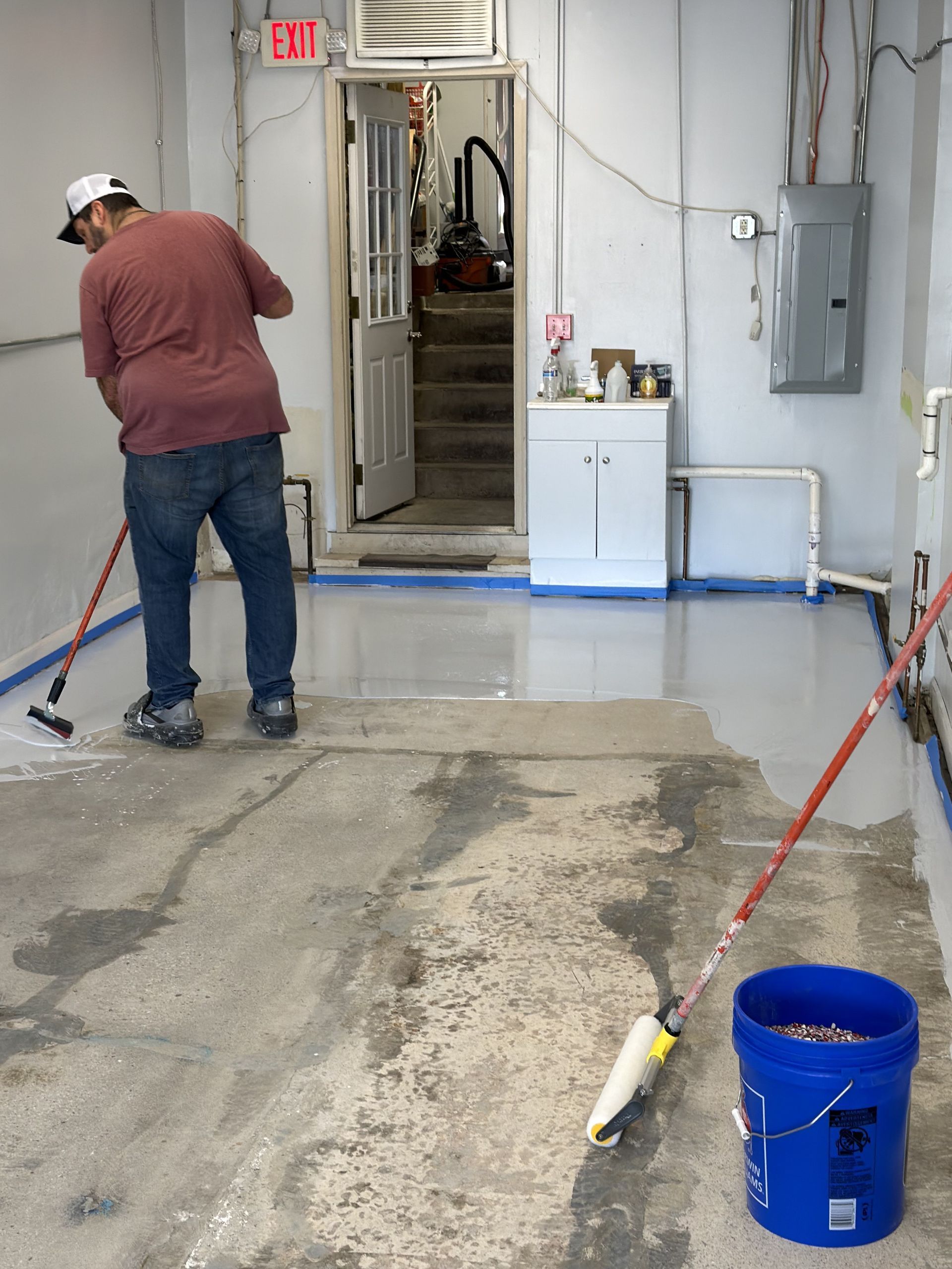A person in a red shirt spreading white floor epoxy with a squeegee in a workshop with unfinished concrete.