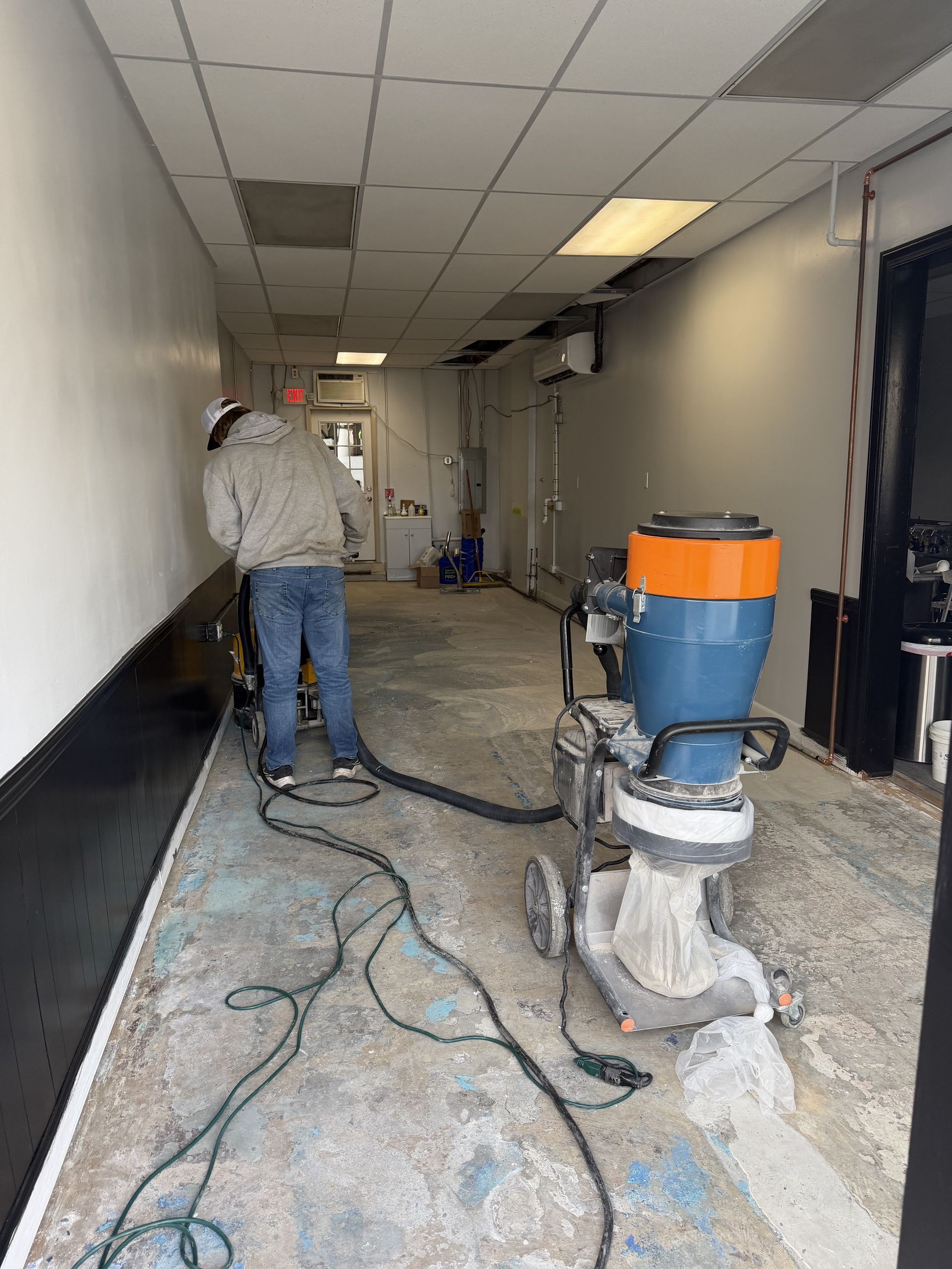 A worker operates a large floor sanding machine in a narrow, unlit commercial space with unfinished walls and floor.