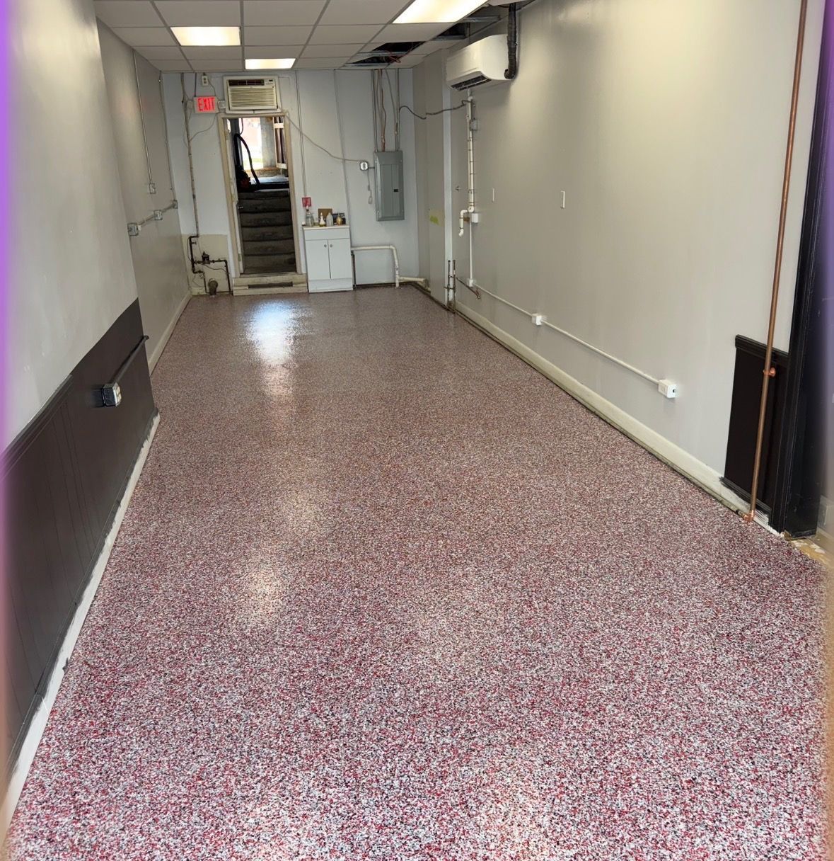 A narrow, empty commercial space with a polished, multicolored speckled floor, white walls, and a back doorway.