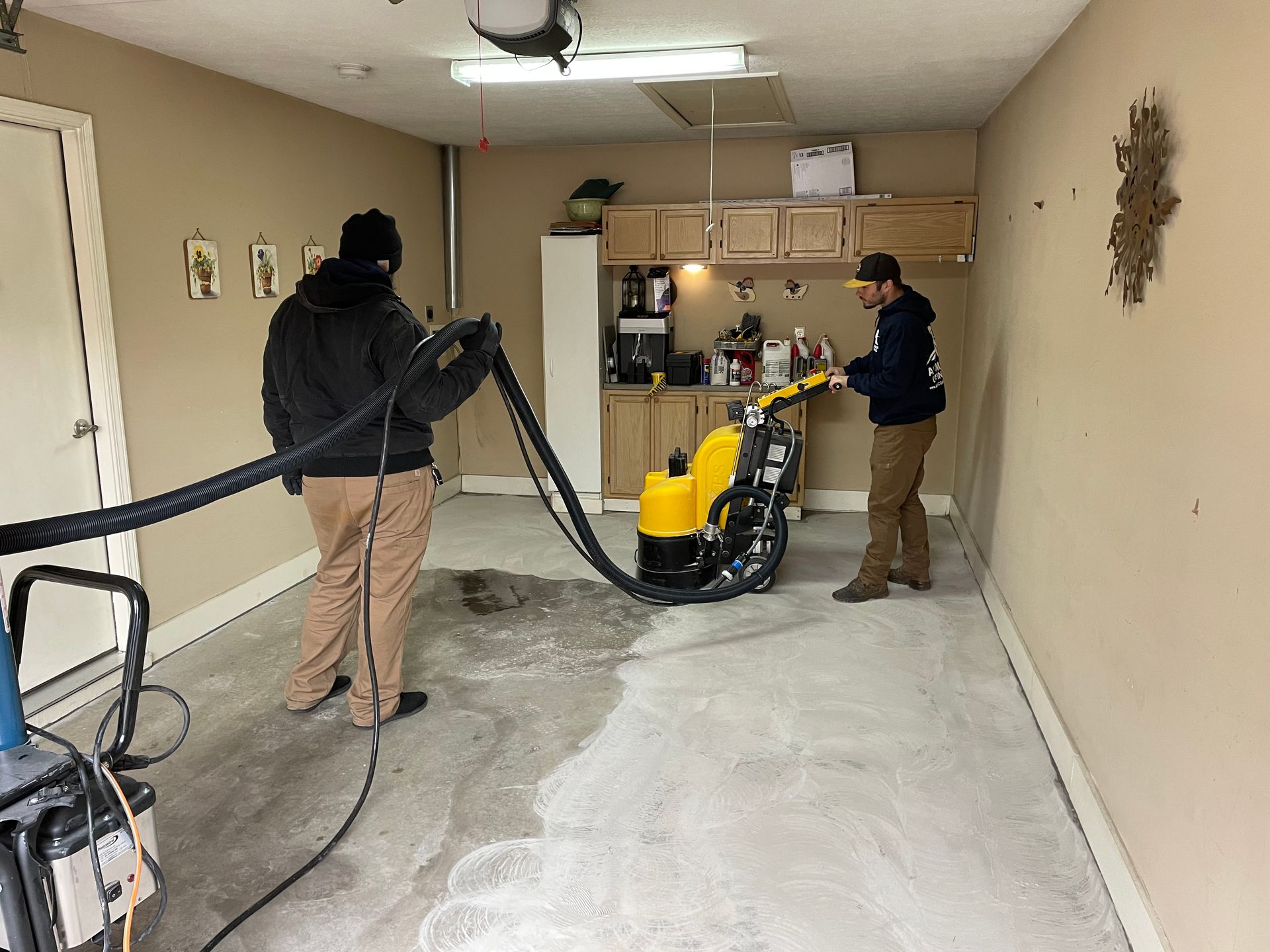 Two people use a floor grinder and industrial vacuum to sand and clean a concrete garage floor.