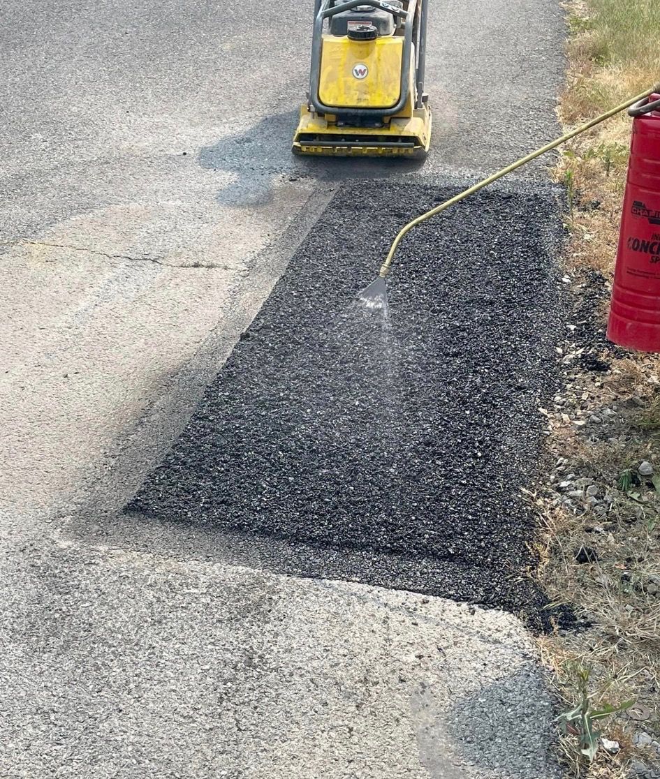 A worker sprays water onto a fresh patch of dark asphalt, with a yellow compactor parked nearby on a paved surface.
