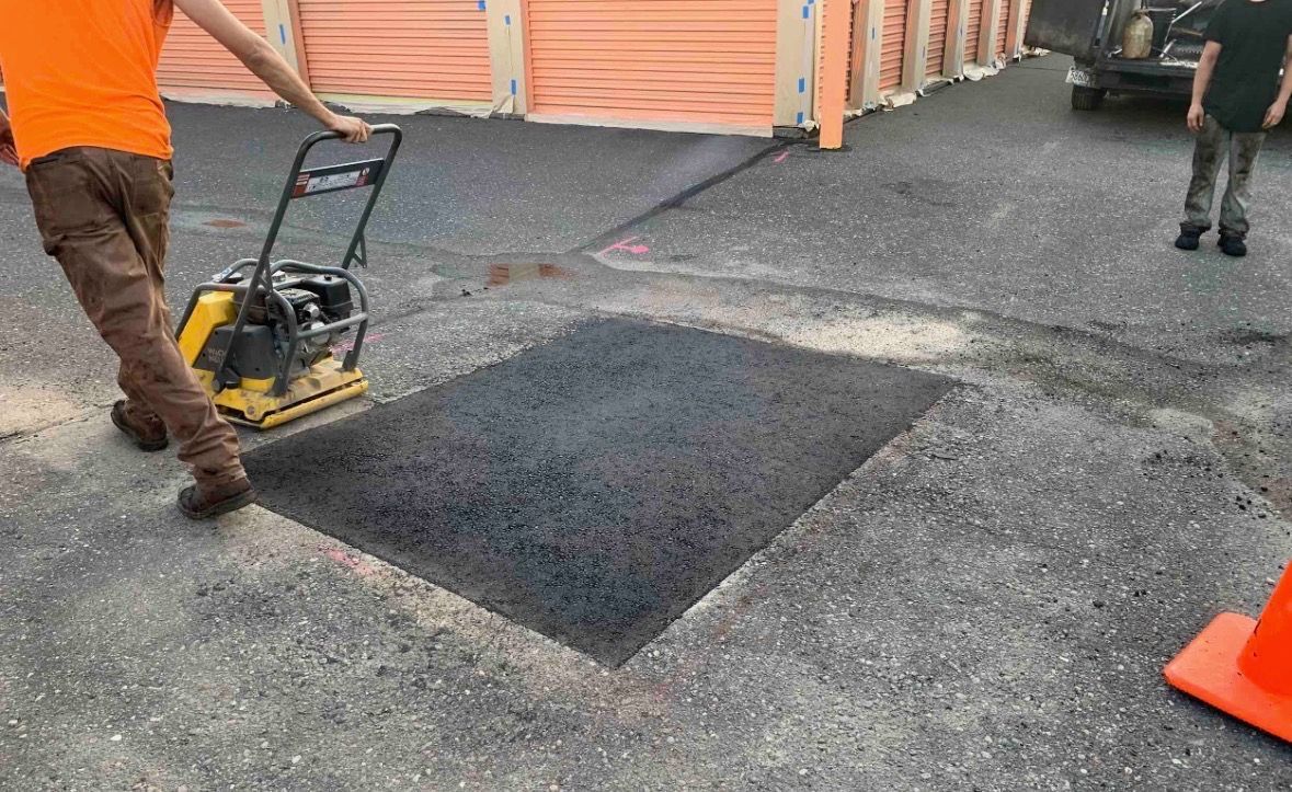 A worker uses a plate compactor on fresh asphalt filling a rectangular patch in a gravel driveway.