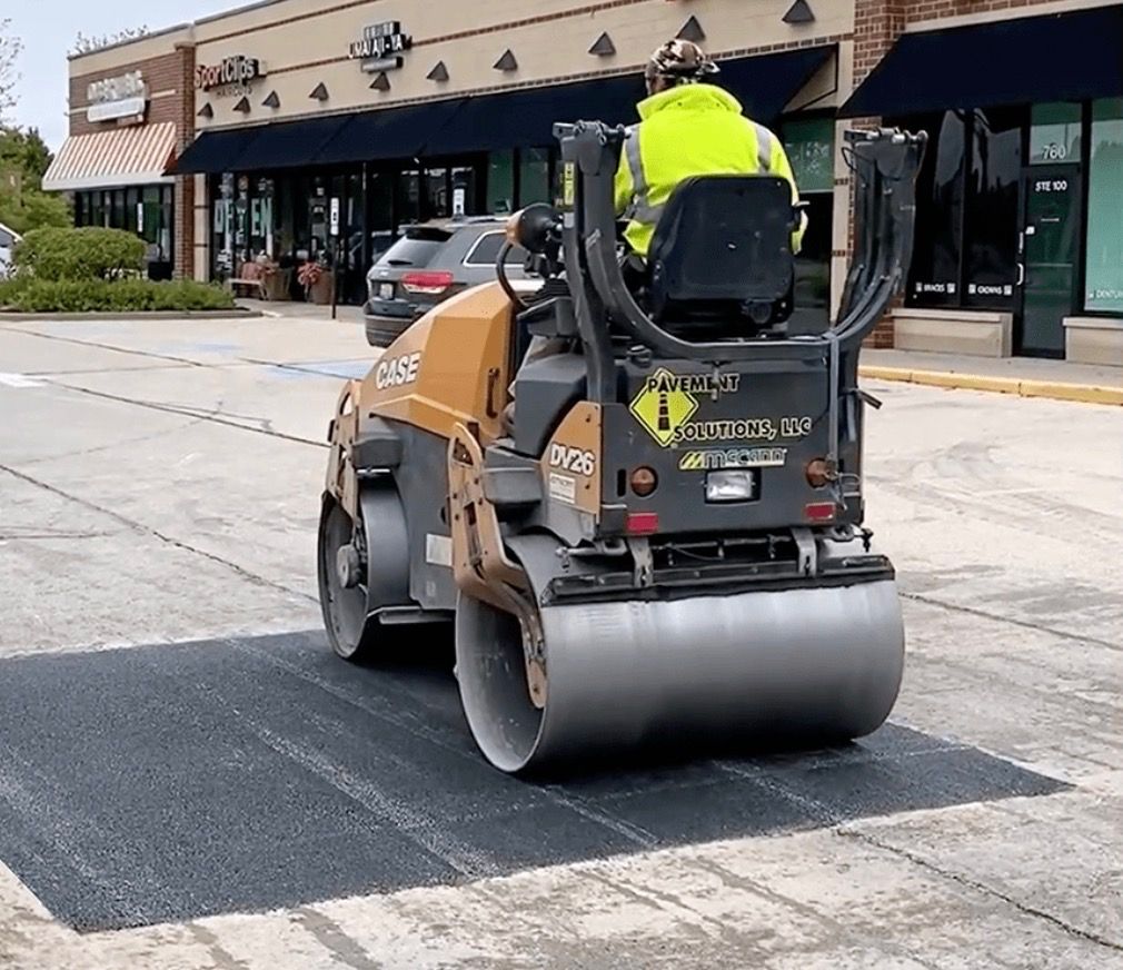 A worker in a bright yellow vest operates a tan construction roller on a patch of fresh asphalt in a commercial parking lot.