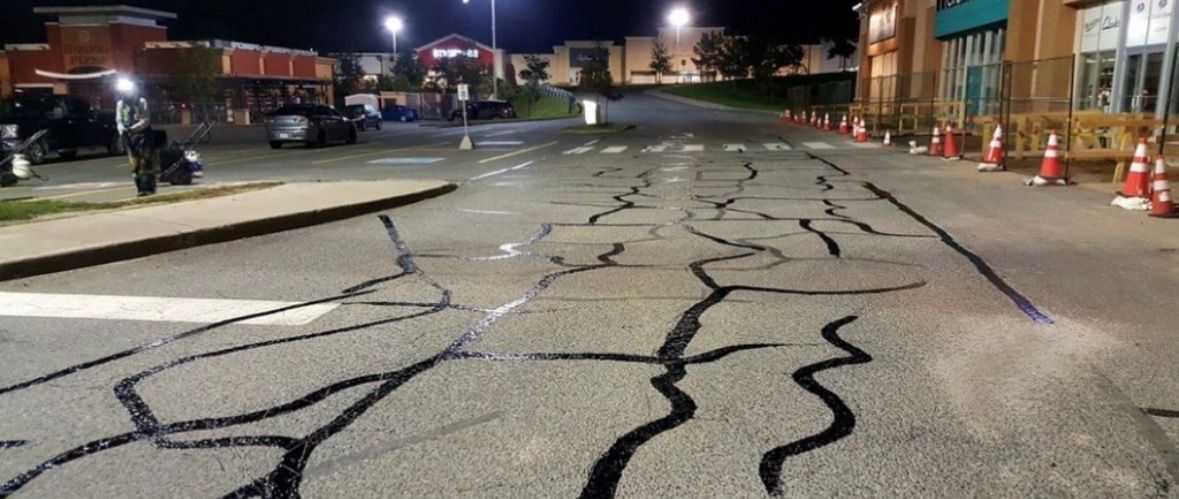 A nighttime view of a parking lot surface marked with black sealant over cracks, near a building with orange traffic cones.
