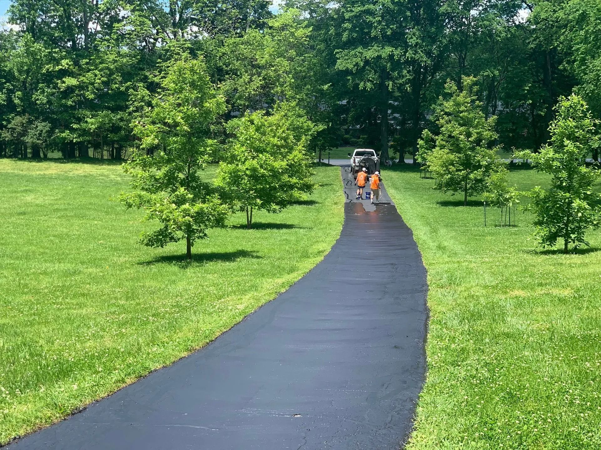 Black asphalt driveway winds through green lawn and trees, with workers in orange vests.