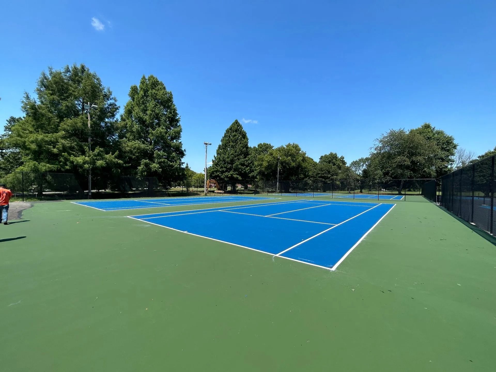 Blue tennis court with white lines, surrounded by green surface, trees, and fence on a sunny day.
