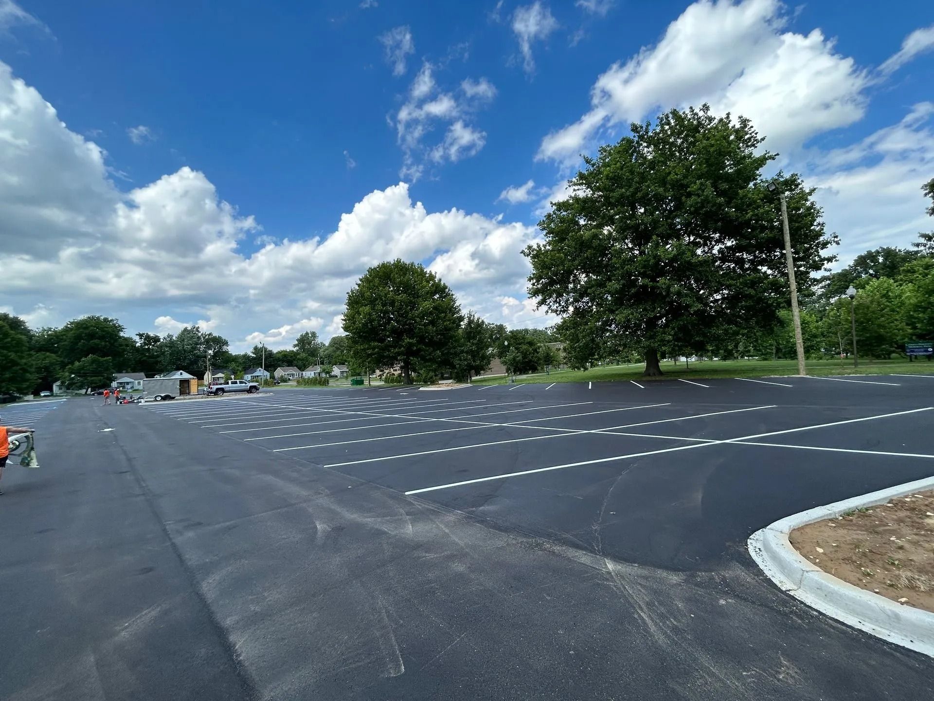 Newly paved parking lot with marked spaces under a blue sky with clouds, trees in the background.