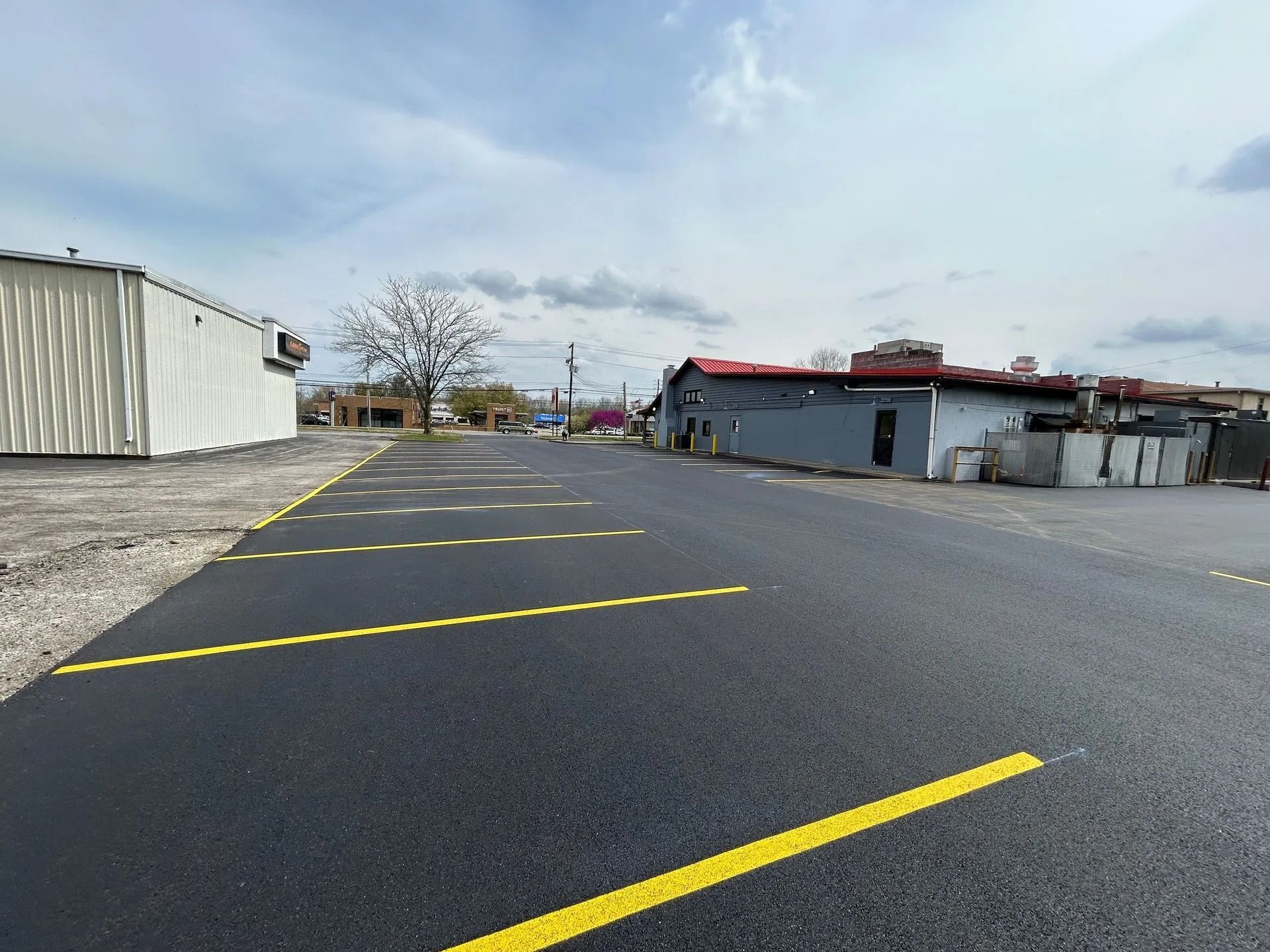 Newly paved asphalt parking lot with yellow painted parking space lines. Buildings are in the background under a cloudy sky.