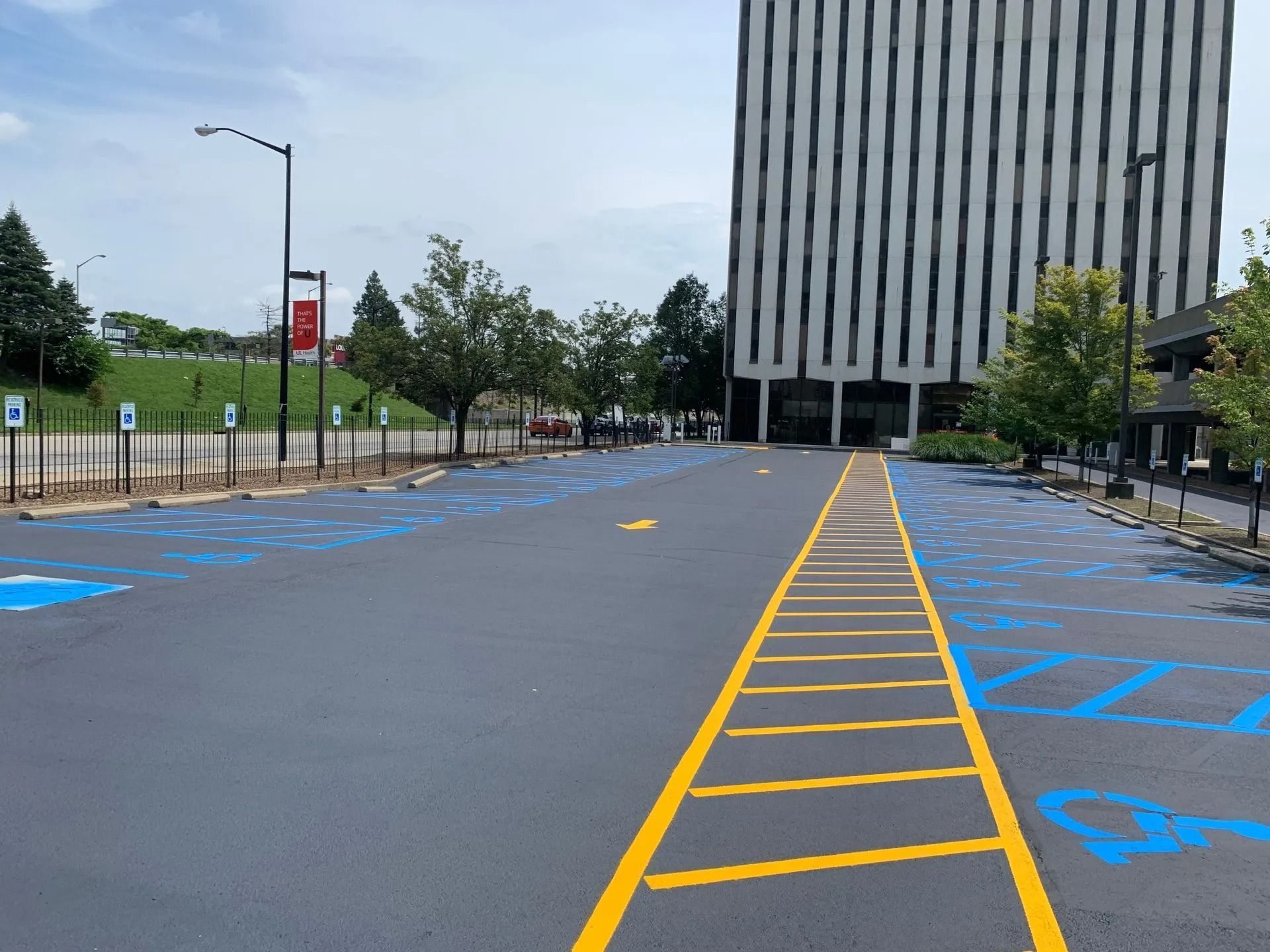 Exterior view of a newly paved parking lot with blue handicap parking spaces, yellow lane markers, and a tall building in the background.