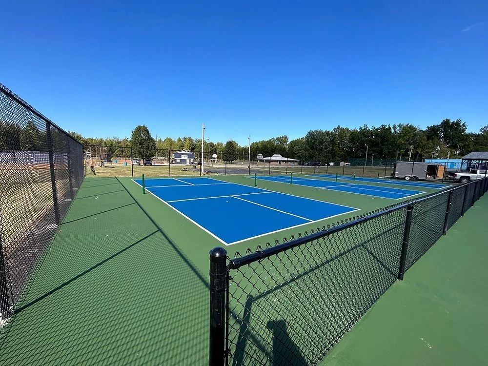 Outdoor pickleball courts with blue playing surfaces and green surrounding areas, enclosed by black fencing.
