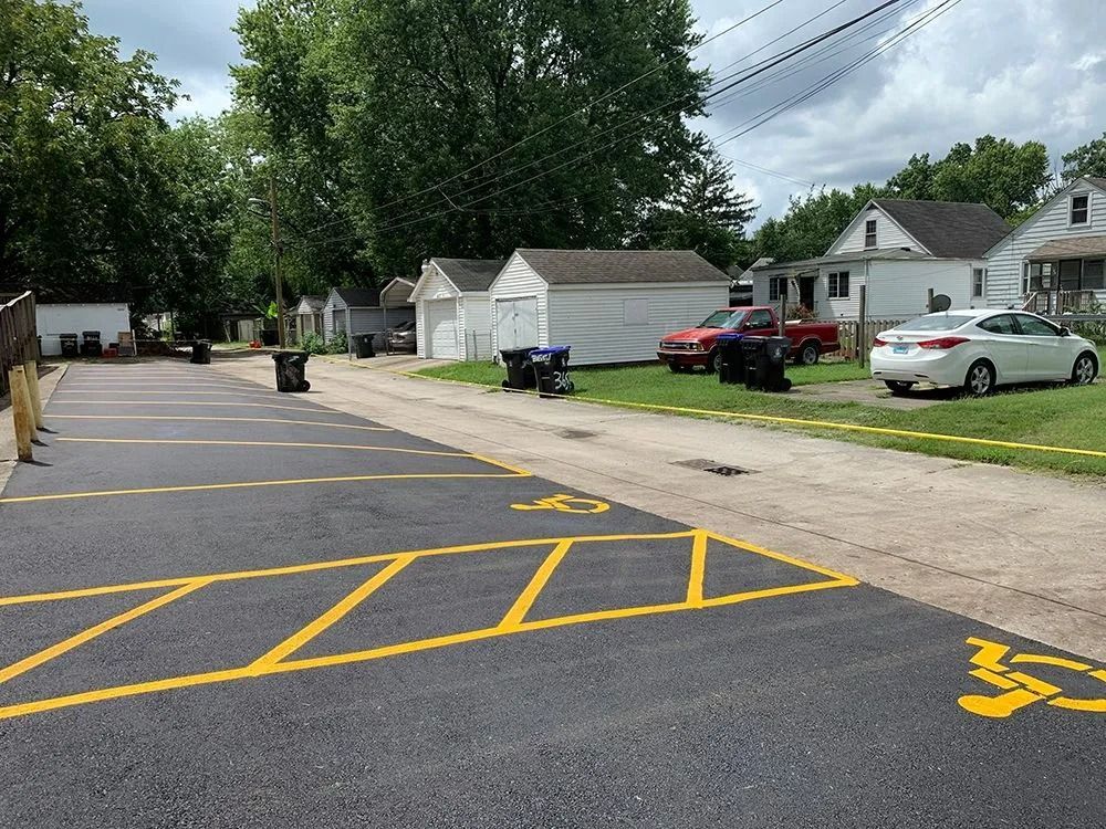 Paved parking lot with handicap symbols. Cars parked near small houses and garages. Trees and sky in background.