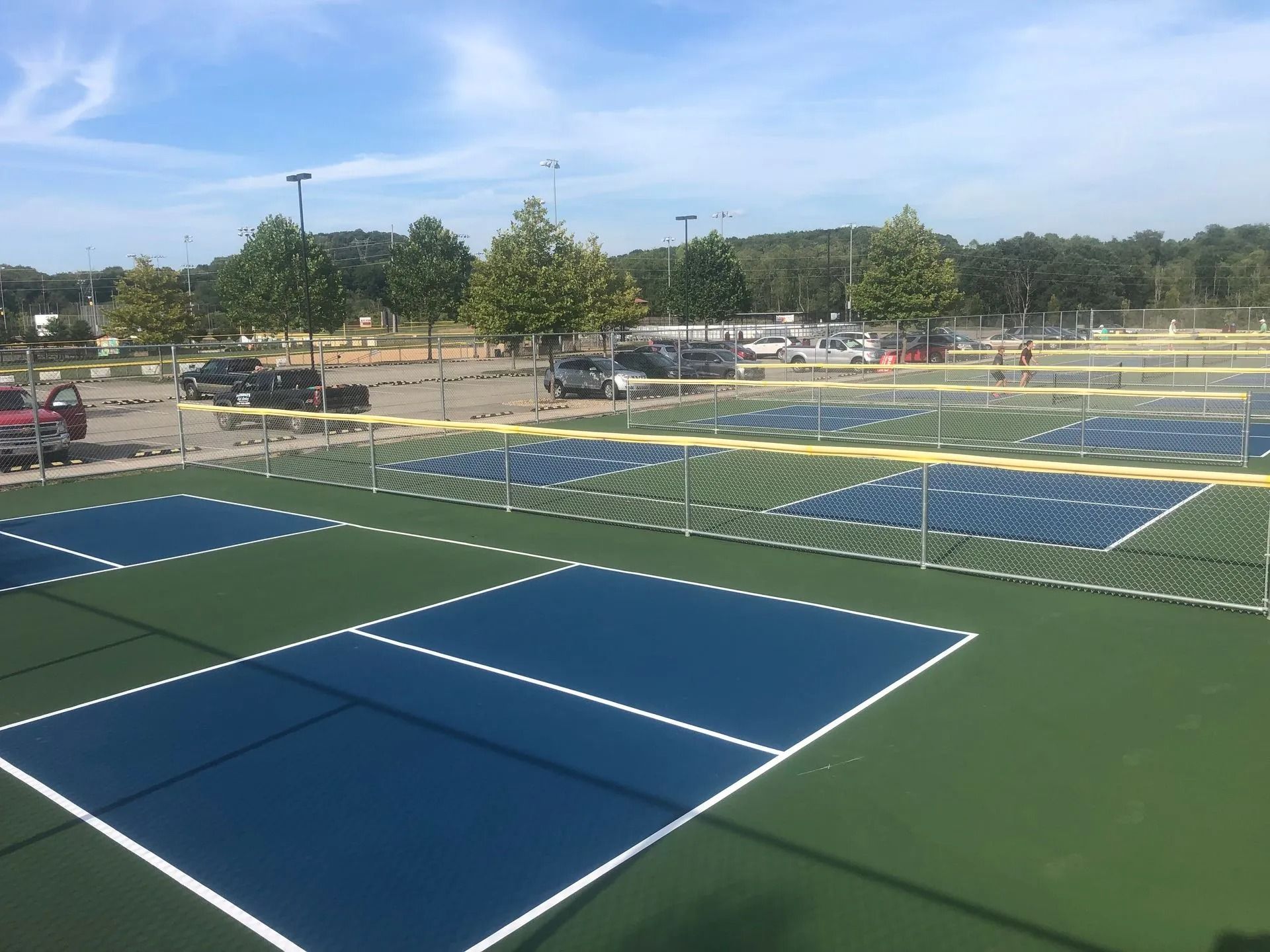 Pickleball courts in an outdoor setting with a parking lot and trees in the background. Courts are blue and green.