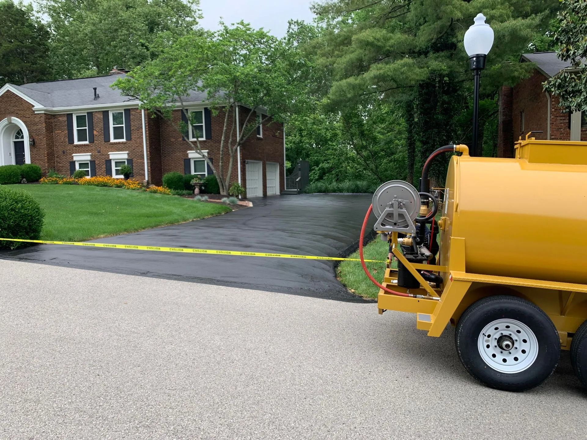 Yellow tanker spraying a black driveway in front of a brick house, yellow caution tape in view.