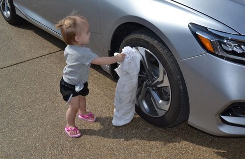 Child with a white towel by the front tire of a silver car on a paved surface.