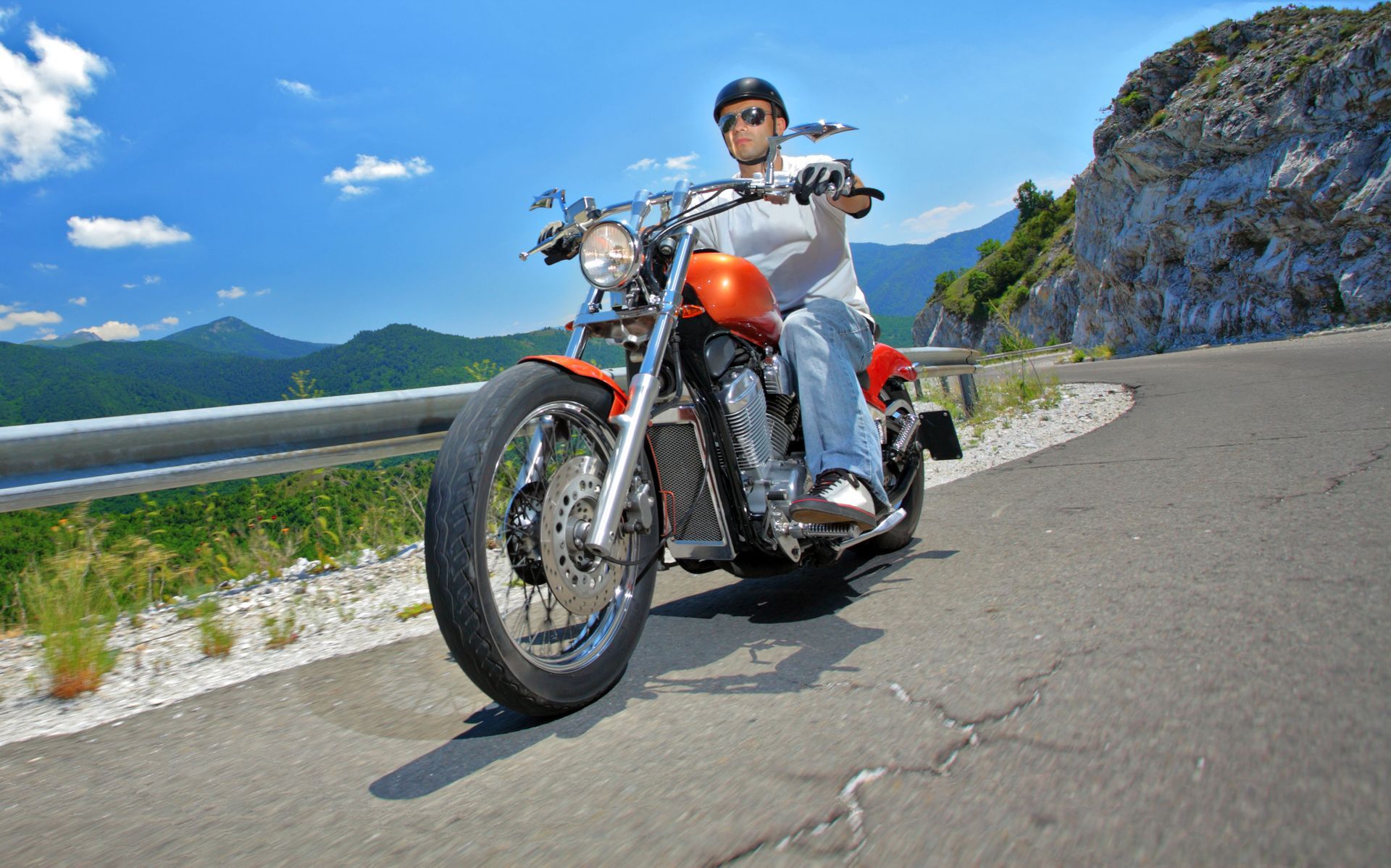 Motorcyclist on an orange motorcycle riding on a winding road with a mountain backdrop under a blue sky.