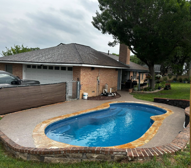 Pool with blue water and tan patio in a backyard with a brick house and tree.