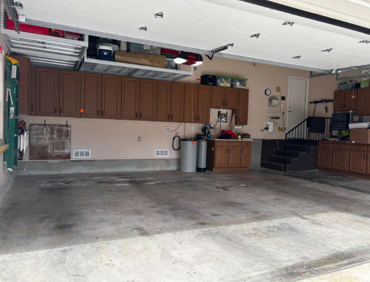 Interior of a garage with brown cabinets, storage shelf, and concrete floor.