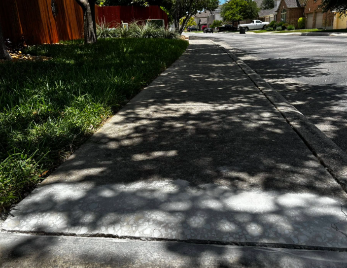 Sidewalk with tree shadows, bordered by grass and a wooden fence. Street and houses in the background.