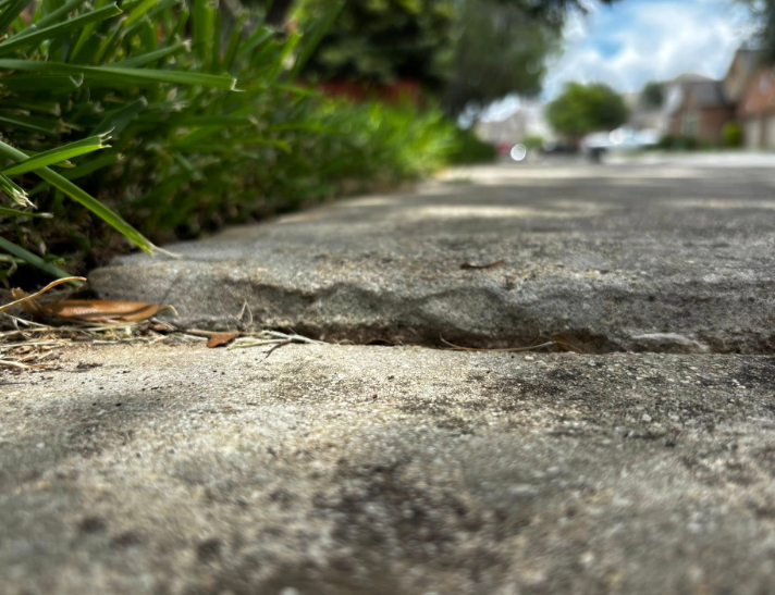 Close-up of cracked concrete sidewalk next to grass, residential street in the background.