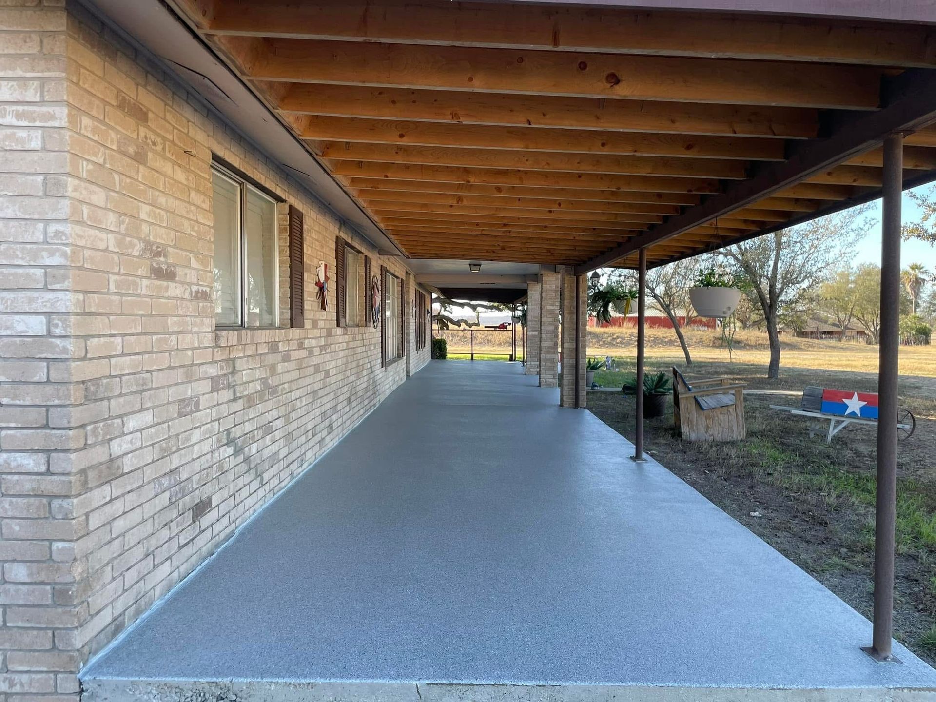 Covered porch with gray speckled flooring, tan brick wall, and brown wooden ceiling.