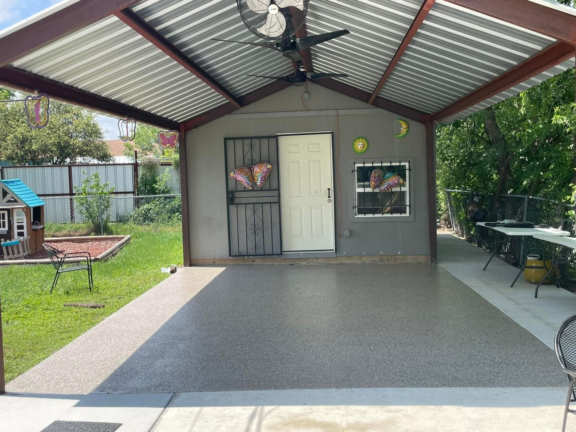 A gray shed with a metal roof and concrete patio in a backyard with a playhouse.