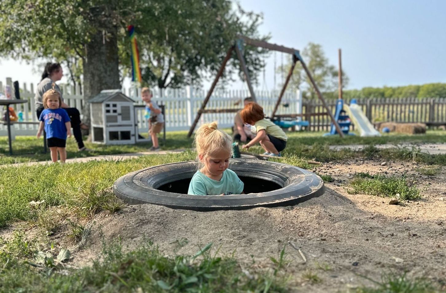 Child in a tire play area, surrounded by other children, swing set, and a fence on a sunny day.