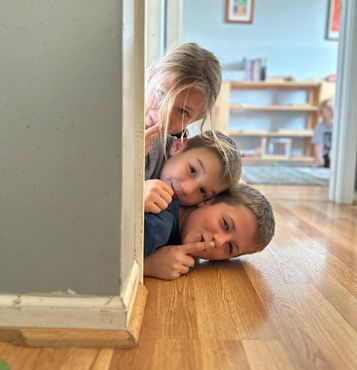 Three children peeking around a doorway, one shushing with a finger to their lips.