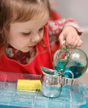 Child pouring blue liquid from a glass pitcher into a silver pitcher, playing with water.