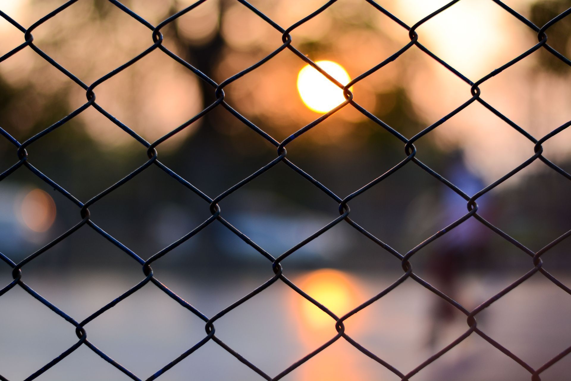 Chain-link fence with a blurred sunset in the background, silhouetting a figure.