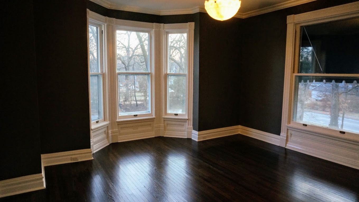 Empty room with dark walls, polished hardwood floors, a bay window, and a white-trimmed window.