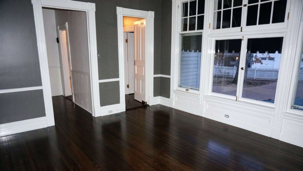 An empty room with dark grey walls, white trim, dark wood floors, and large windows looking out into the night.