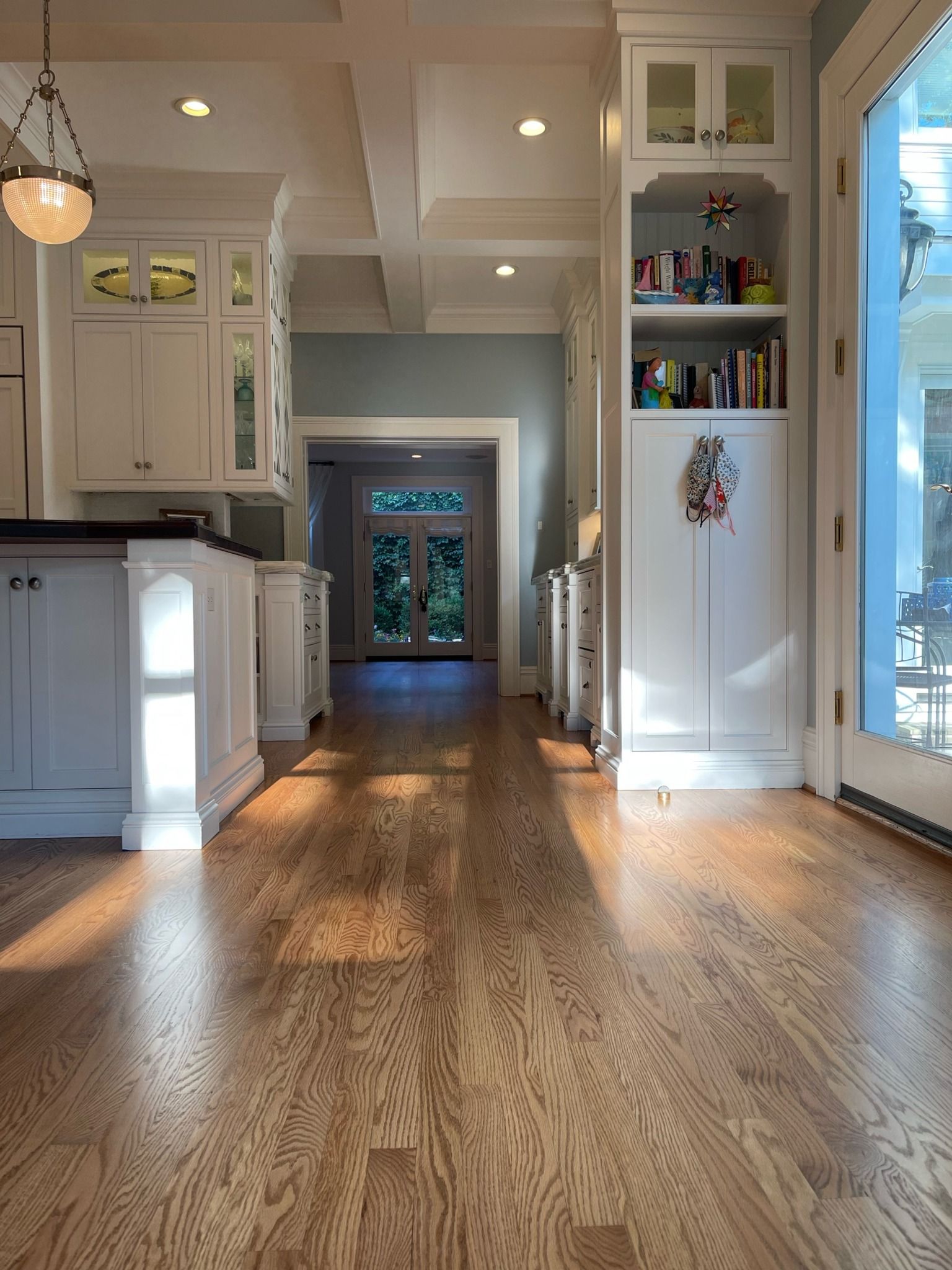 A low-angle view of a bright kitchen with light oak floors, white cabinetry, a built-in bookcase, and a distant doorway.
