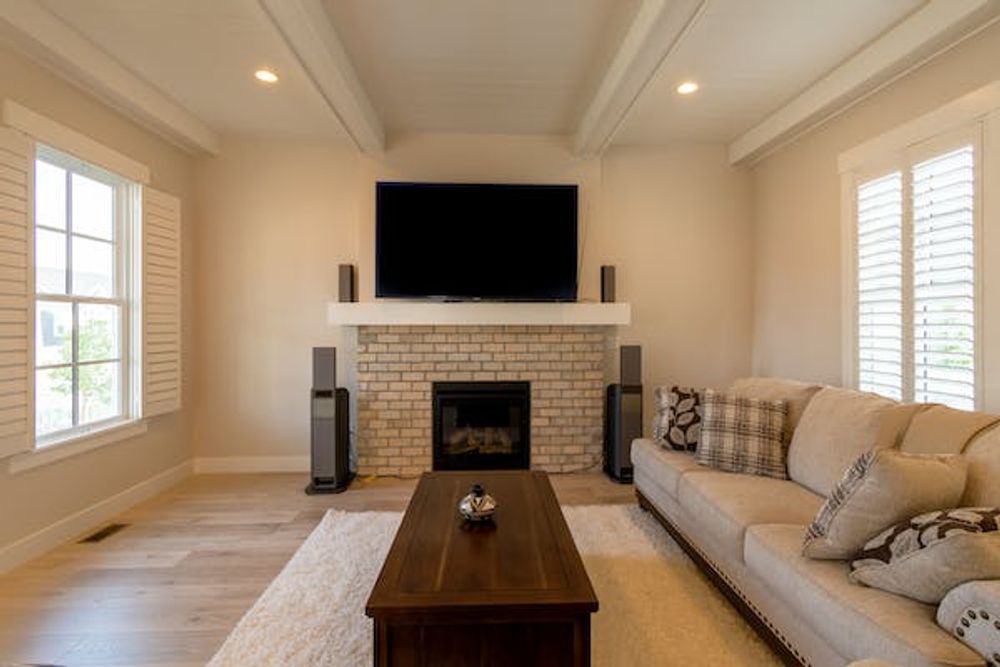 A living room with a beige couch, a brick fireplace with a wall-mounted TV, a coffee table, and plantation shutters.
