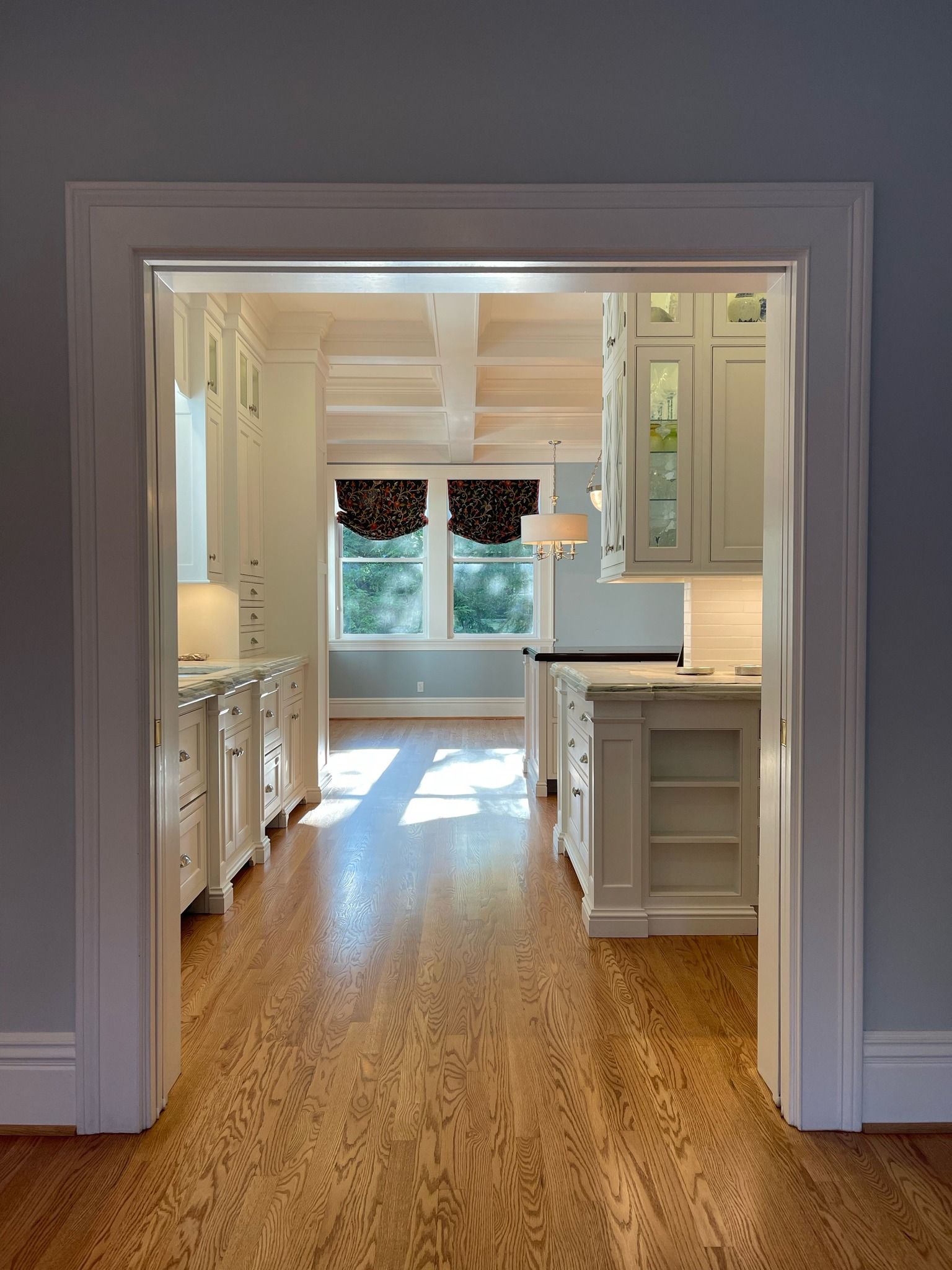 A view through a white door frame into a bright, modern white kitchen with wood floors and windows at the far end.