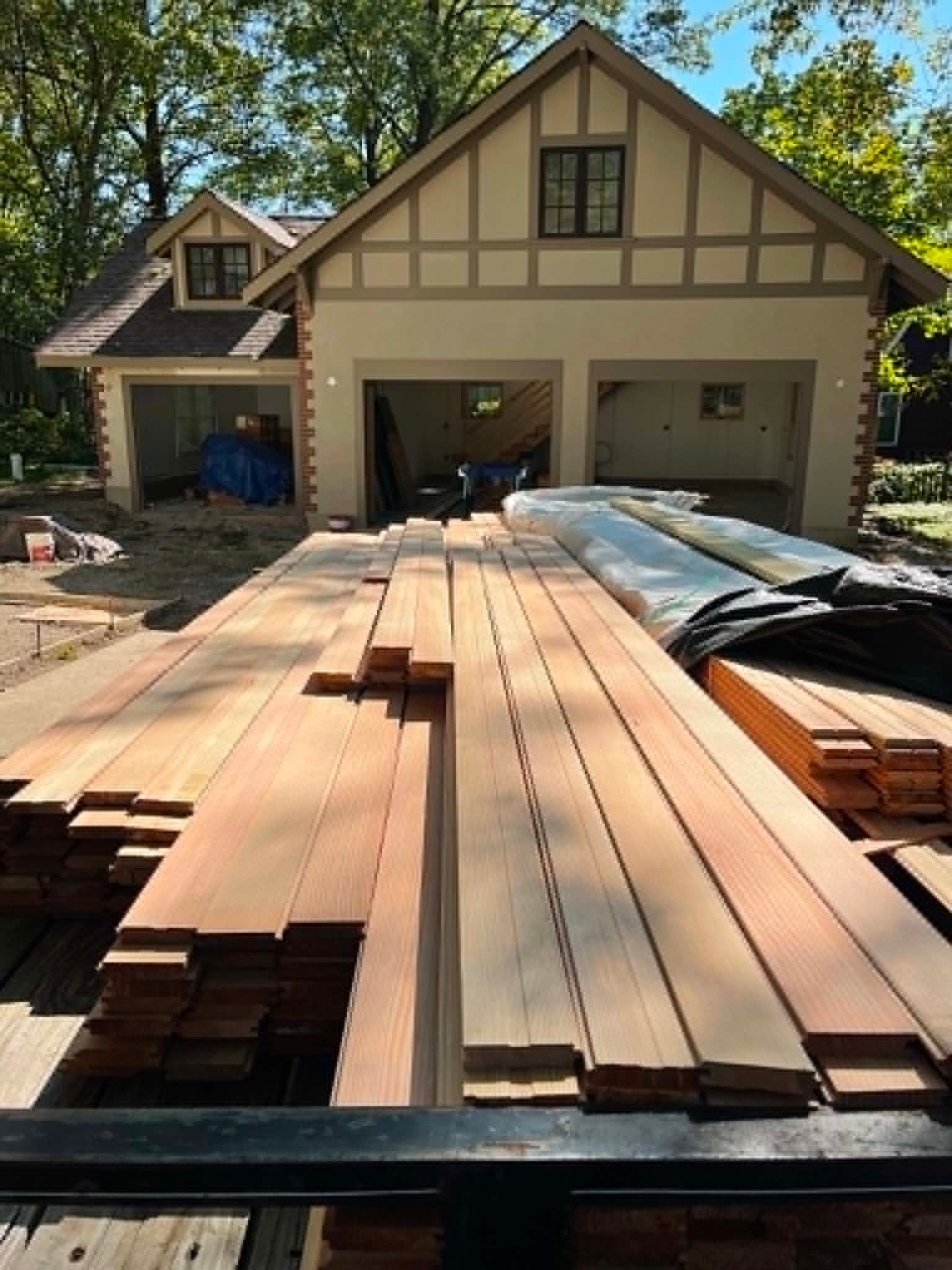Stacked lumber on a driveway in front of a house under construction, with an open garage and driveway tarps.