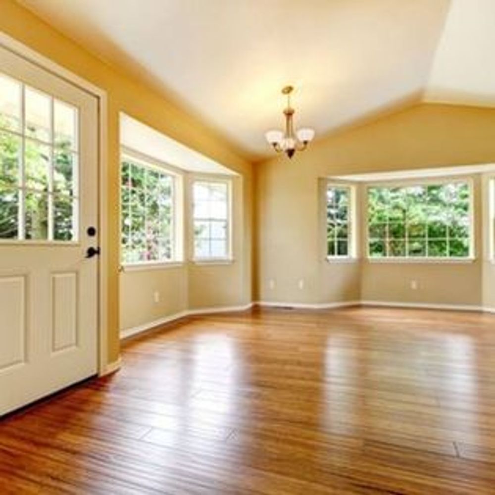 An empty room with light wood floors, beige walls, large windows, and a hanging chandelier near a white door.
