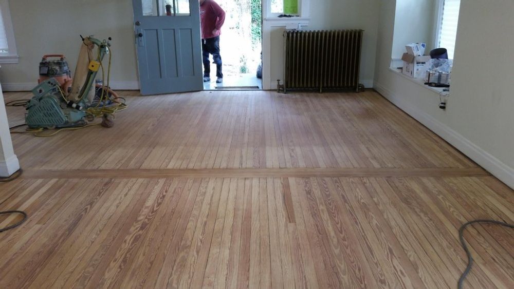A person operates a floor sanding machine in a room with light-toned, newly sanded wooden floors and a blue door.