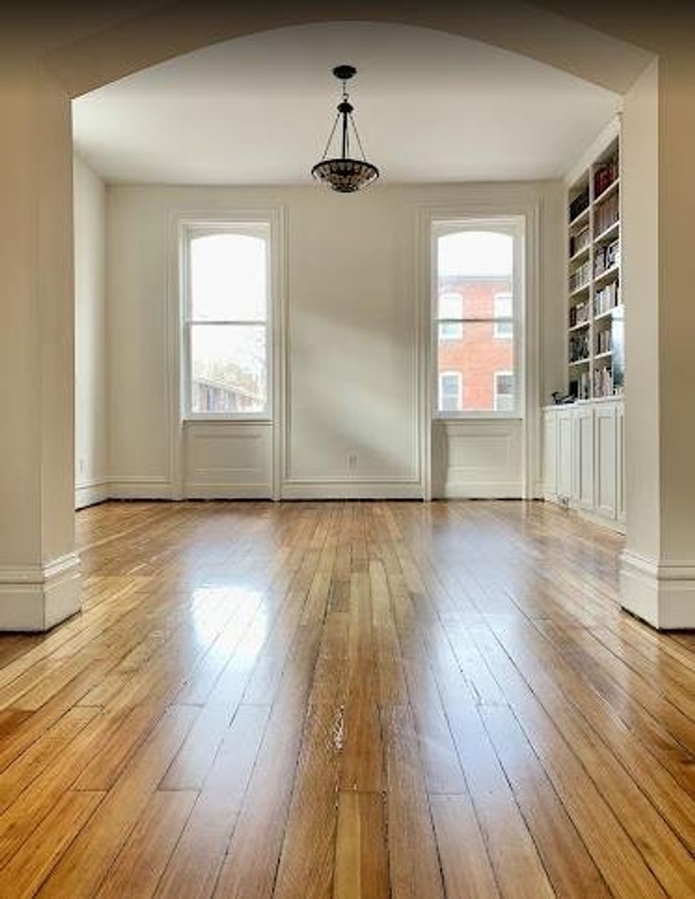A bright, empty room with polished hardwood floors, two windows, a ceiling light, and a built-in bookshelf.
