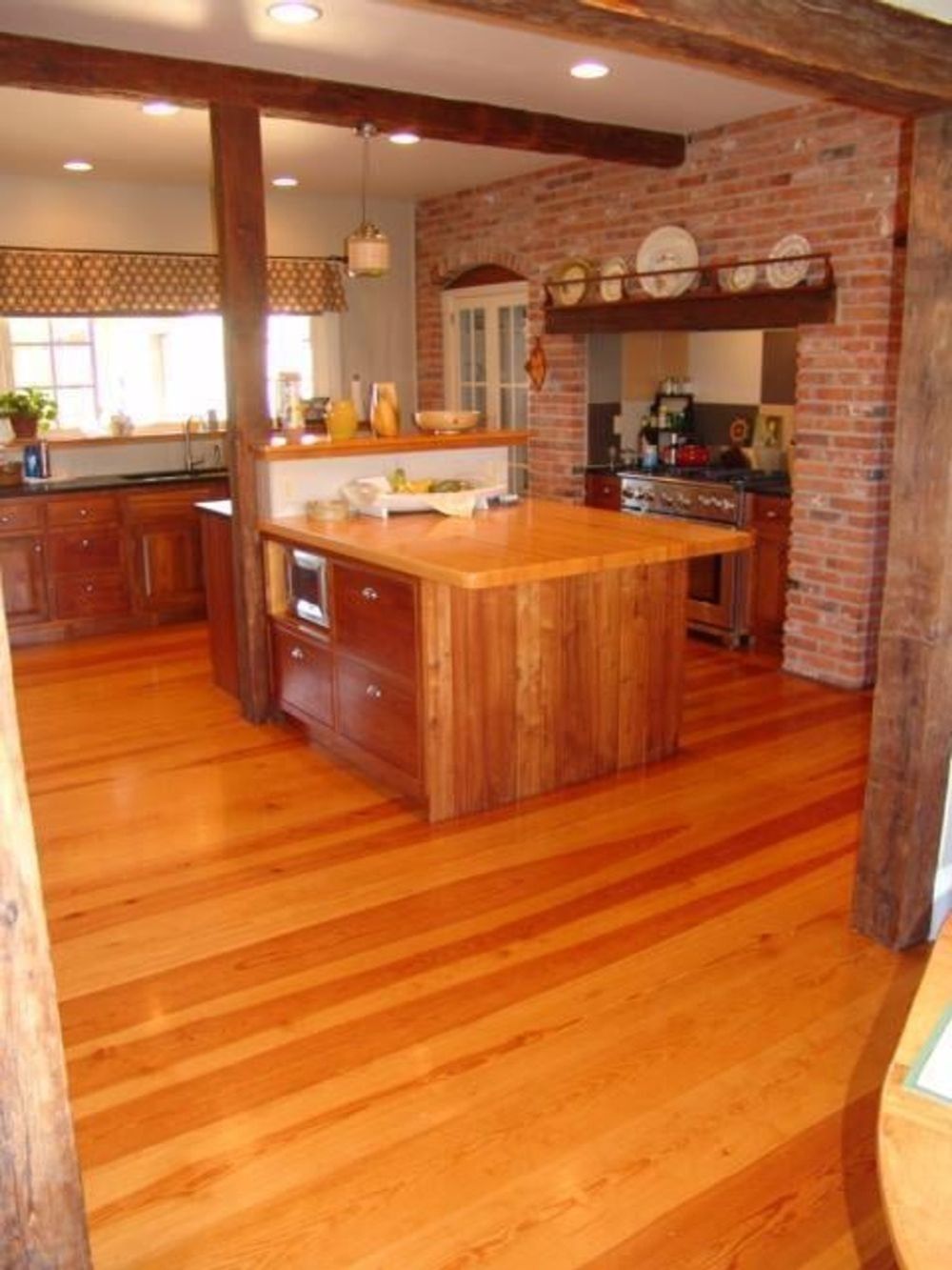 A rustic kitchen with wood floors, a wooden center island, brick wall, and exposed timber ceiling beams.